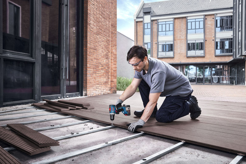 Eine Person mit Sicherheitsausrüstung verwendet eine Akku-Bohrmaschine, um Terrassendielen im Außenbereich zu installieren. Anwendungsbild