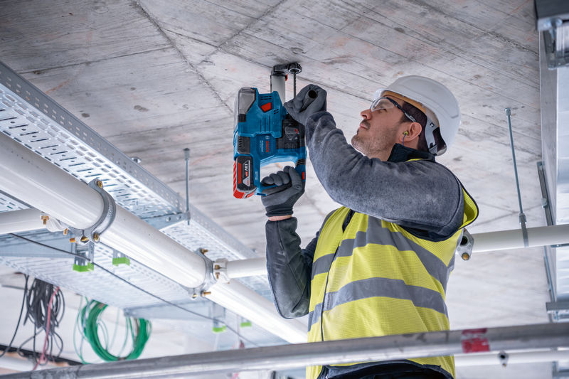 Eine Person mit Sicherheitsausrüstung bohrt auf einer Baustelle in eine Betondecke. Anwendungsbild