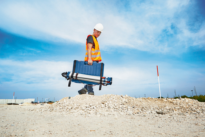 Eine Person mit Sicherheitsausrüstung trägt einen Koffer und ein Stativ über eine Baustelle. Anwendungsbild