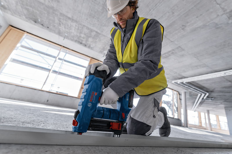 Eine Person mit Sicherheitsausrüstung verwendet einen kabellosen Betonnagler auf einer Metallschiene. Anwendungsbild