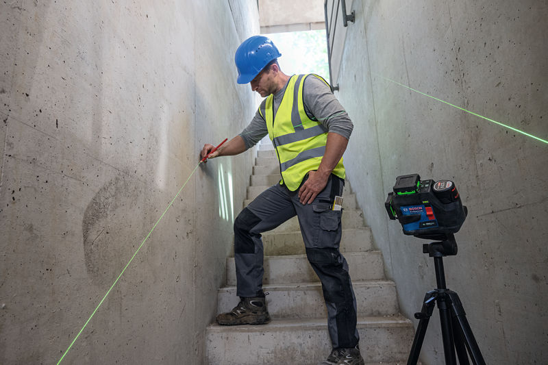 Eine Person mit Sicherheitsausrüstung markiert eine Wand mit einem Lasernivelliergerät auf einem Stativ. Anwendungsbild
