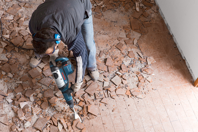 Eine Person mit Schutzausrüstung zerschlägt Bodenfliesen mit einem Abbruchhammer. Anwendungsbild