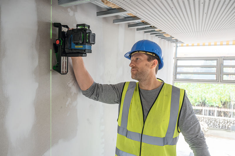 Eine Person mit Sicherheitsausrüstung positioniert ein Lasernivelliergerät an einer Wand. Anwendungsbild