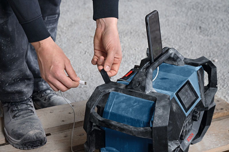Eine Person steckt ein Ladekabel in ein robustes Baustellenradio, an dem ein Telefon angedockt ist. Anwendungsbild