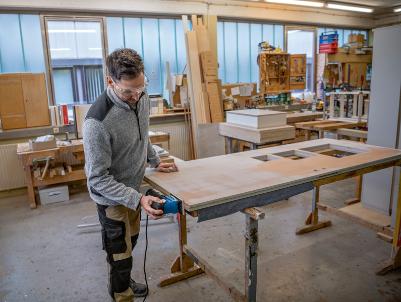 Eine Person mit Schutzausrüstung schleift in einer Werkstatt eine Holztür mit einem Schwingschleifer. Anwendungsbild
