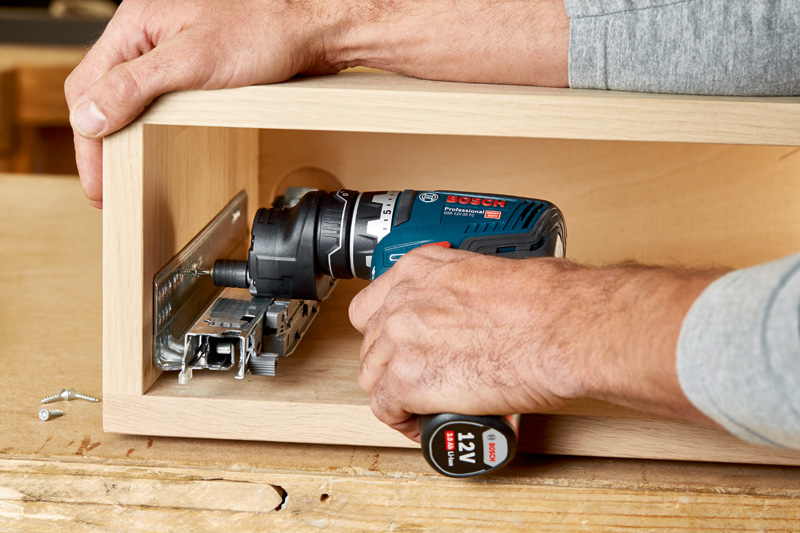 Person, die mit einer Akkubohrmaschine eine Schubladenführung aus Metall in einer Holzkiste installiert. Anwendungsbild