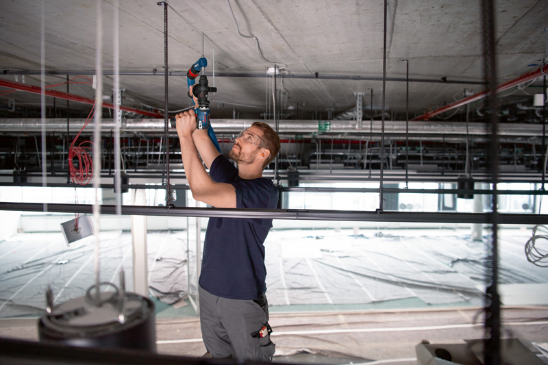Eine Person mit Schutzausrüstung bohrt mit einem Elektrowerkzeug mit FlexiClick-Adapter in eine Betondecke. Anwendungsbild