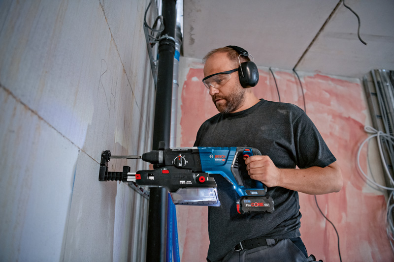 Eine Person mit Schutzausrüstung bohrt mit einem Werkzeug mit integrierter Staubabsaugung in eine Wand. Anwendungsbild