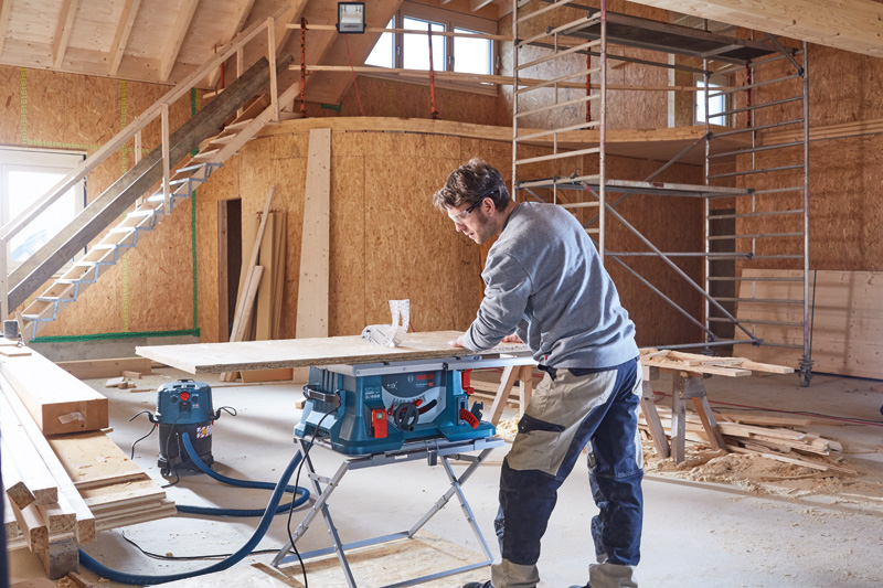 Eine Person bedient eine Tischkreissäge, um in einem teilweise fertiggestellten Gebäude Holz zu schneiden. Anwendungsbild