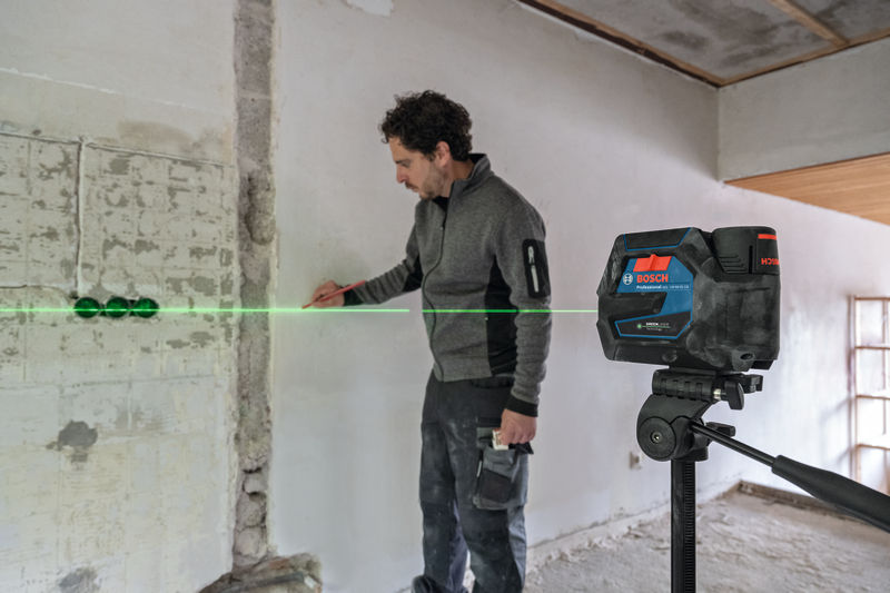 Eine Person markiert eine Wand mit einem auf einem Stativ montierten Lasernivelliergerät. Anwendungsbild