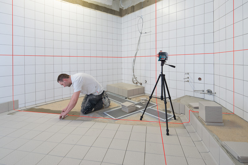 Eine Person verlegt Bodenfliesen in einem Badezimmer, wobei sie sich dabei von einem Lasernivelliergerät auf einem Stativ leiten lässt. Anwendungsbild