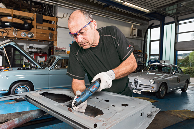 Eine Person mit Sicherheitsausrüstung verwendet ein kabelloses Rotationswerkzeug zum Schleifen von Metall in einer Garage. Anwendungsbild