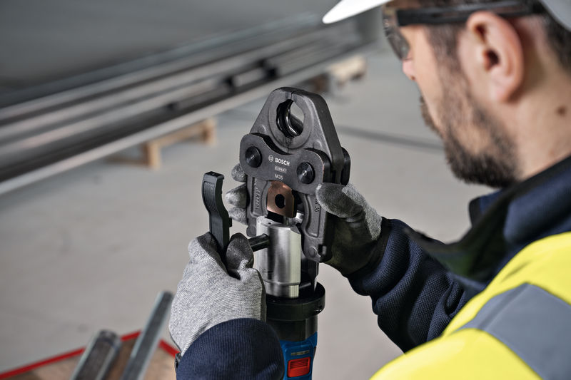Eine Person mit Schutzausrüstung bedient ein Akku-Presswerkzeug an einer Metallarmatur. Anwendungsbild
