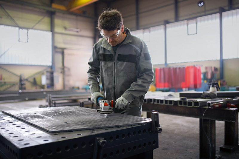 Eine Person mit Schutzausrüstung verwendet einen Winkelschleifer auf einem Metallarbeitstisch. Anwendungsbild