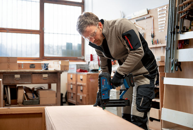 Eine Person mit Sicherheitsausrüstung verwendet einen Akku-Holznagler auf einem Holzbrett. Anwendungsbild