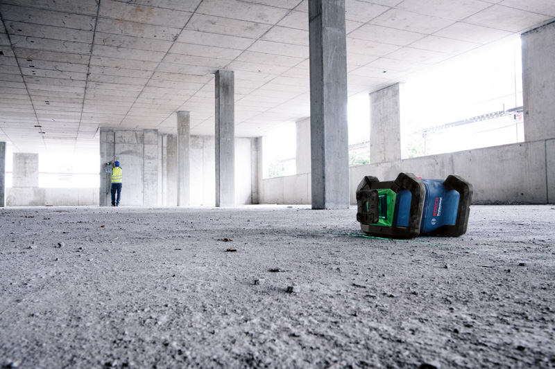 Lasernivellierungsgerät auf einem Betonboden in einem großen Raum platziert, Person trägt Sicherheitsausrüstung. Anwendungsbild