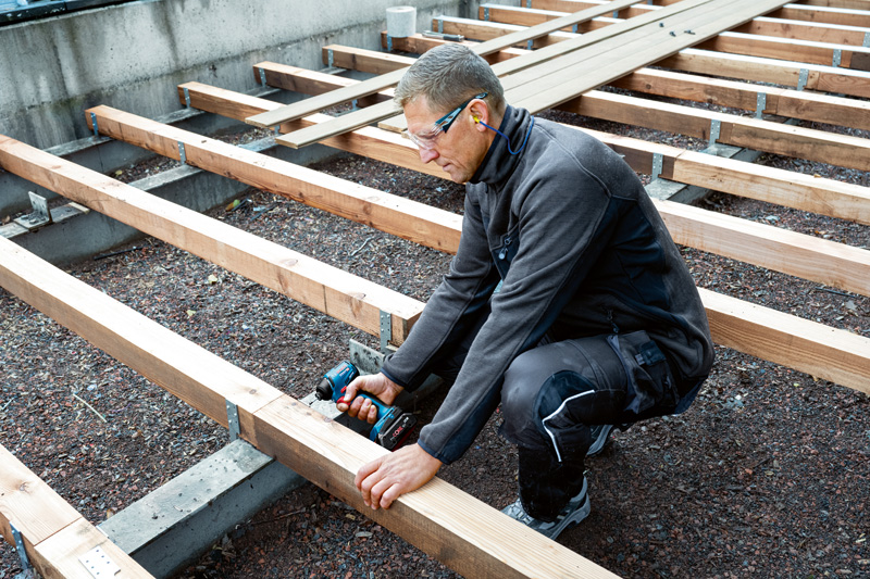 Eine Person mit Sicherheitsausrüstung befestigt auf einer Baustelle Holzbalken mit einer Bohrmaschine. Anwendungsbild