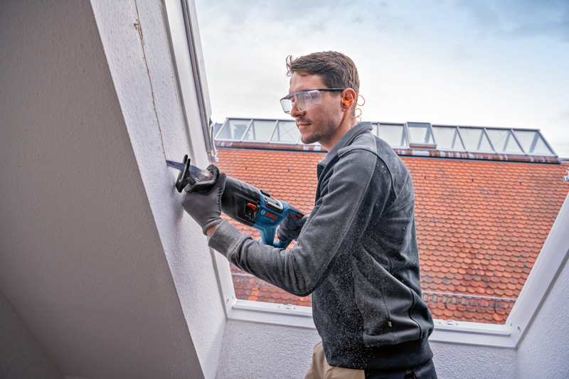 Eine Person mit Schutzausrüstung schneidet mit einer Säbelsäge in eine Wand. Anwendungsbild