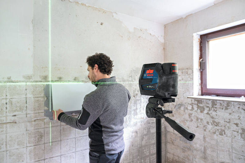 Eine Person richtet Wandfliesen in einem teilweise renovierten Raum mit einem Lasernivelliergerät aus. Anwendungsbild