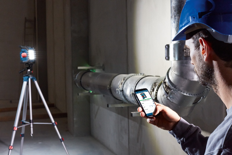 Eine Person mit Sicherheitsausrüstung verwendet ein Smartphone in der Nähe eines Lasernivelliergeräts und von Metallrohren. Anwendungsbild