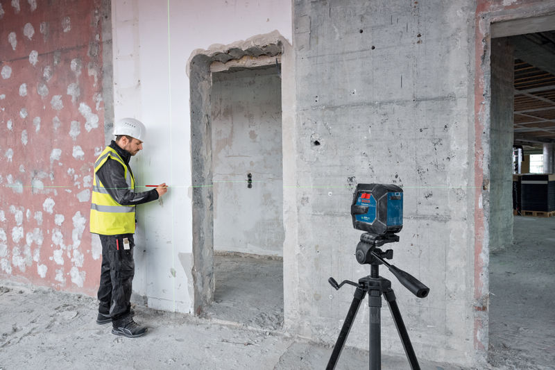Eine Person mit Sicherheitsausrüstung markiert eine Wand mit einem Lasernivelliergerät auf einem Stativ. Anwendungsbild