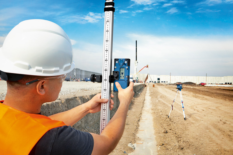 Eine Person mit Sicherheitsausrüstung hält auf einer Baustelle einen Laserempfänger an eine Nivellierlatte. Anwendungsbild
