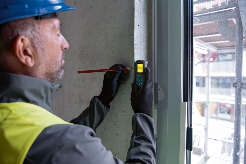 Eine Person mit Sicherheitsausrüstung markiert mit einem Lasermessgerät eine Stelle auf einer Betonwand. Anwendungsbild