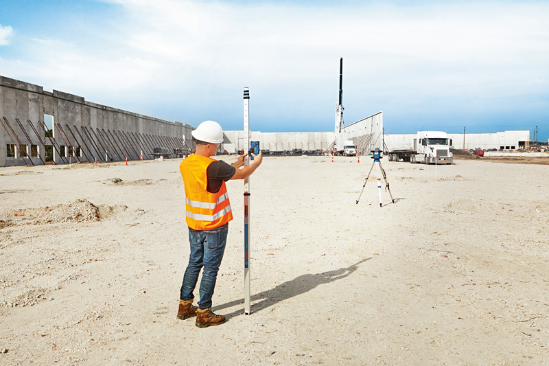 Eine Person mit Sicherheitsausrüstung verwendet auf einer Baustelle ein Lasernivelliergerät mit Laserempfänger. Anwendungsbild