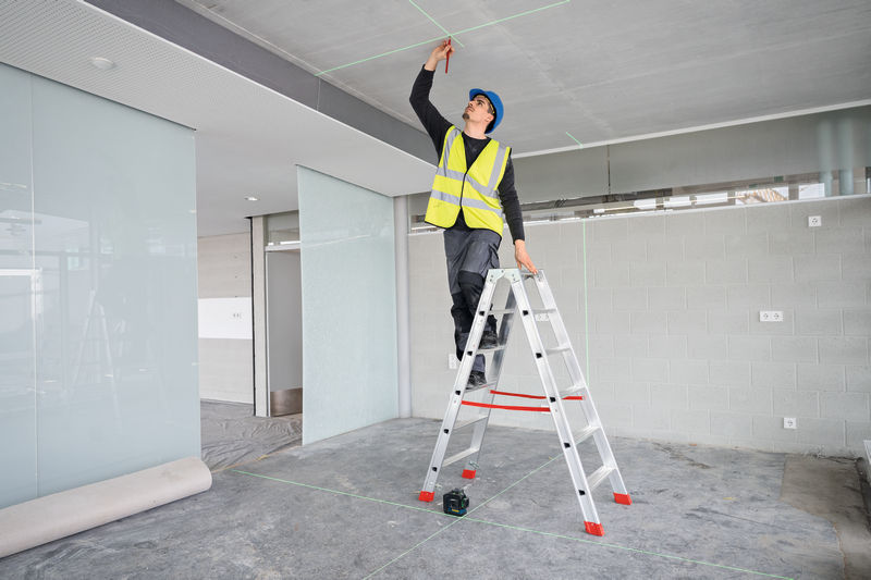 Eine Person mit Sicherheitsausrüstung markiert eine Decke mit einem Lasernivelliergerät. Anwendungsbild