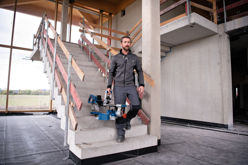 Eine Person mit einer Akku-Gehrungssäge geht auf einer Baustelle die Treppe hinunter. Anwendungsbild