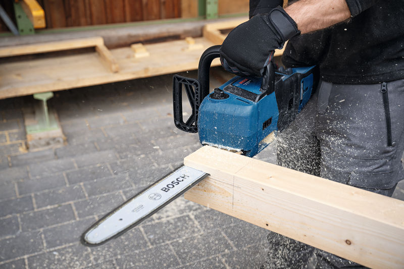 Eine Person mit Sicherheitsausrüstung schneidet mit einer blauen Kettensäge einen Holzbalken durch. Anwendungsbild Nahaufnahme