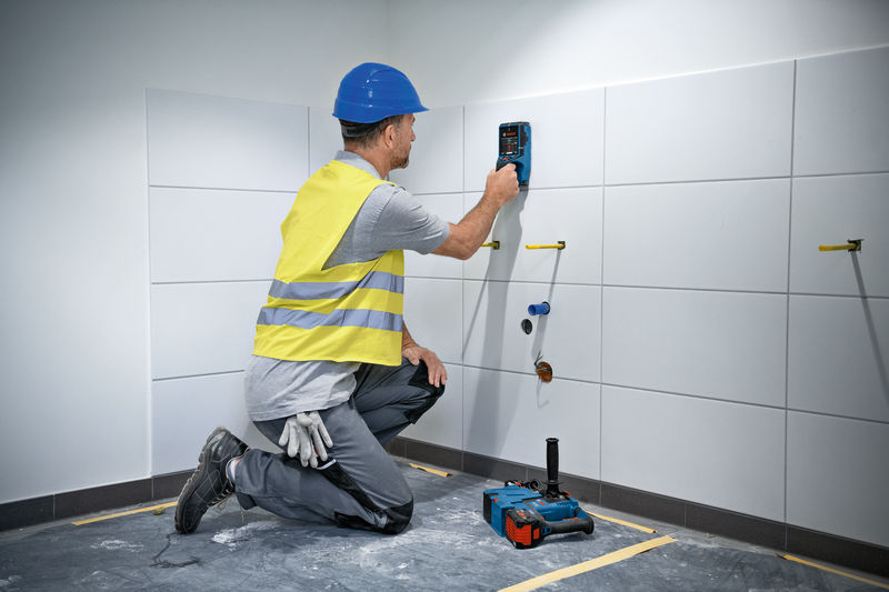 Eine Person mit Schutzausrüstung verwendet einen Wandscanner an einer gefliesten Wand. Anwendungsbild
