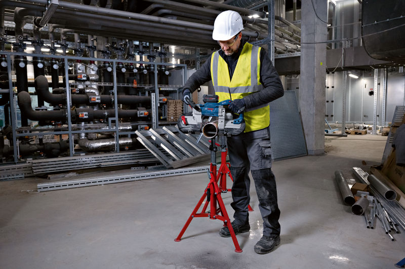 Eine Person mit Sicherheitsausrüstung schneidet ein Metallrohr mit einer Akku-Bandsäge auf einem Stativ. Anwendungsbild