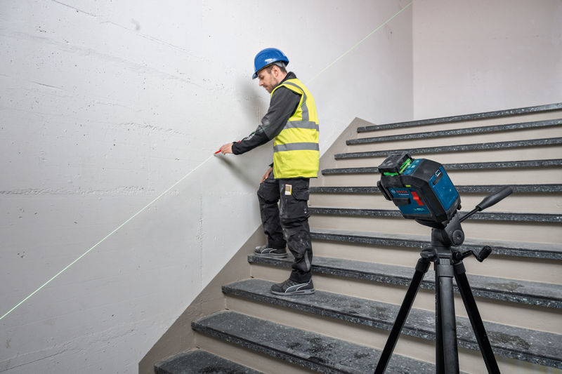 Eine Person mit Sicherheitsausrüstung markiert eine Wand mit einem Lasernivelliergerät auf einem Stativ. Anwendungsbild