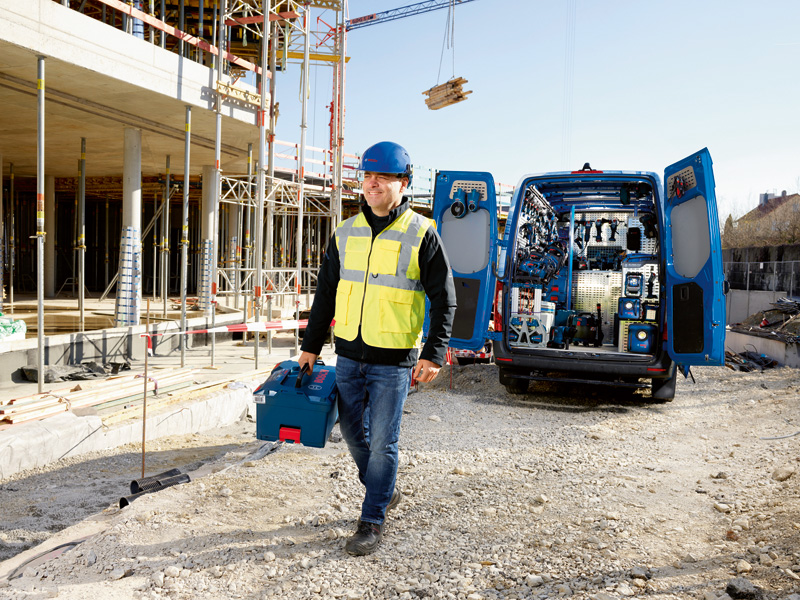Ein Arbeiter in Sicherheitsausrüstung trägt einen Werkzeugkoffer neben einem mit Elektrowerkzeugen vollgestopften Lieferwagen. Anwendungsbild