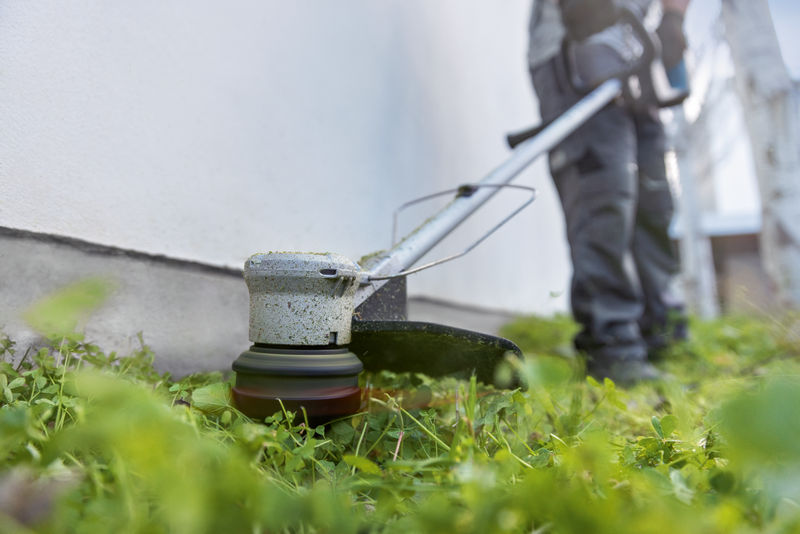 Eine Person mit Sicherheitsausrüstung schneidet mit einem Akku-Rasentrimmer Gras entlang einer Wand. Anwendungsbild