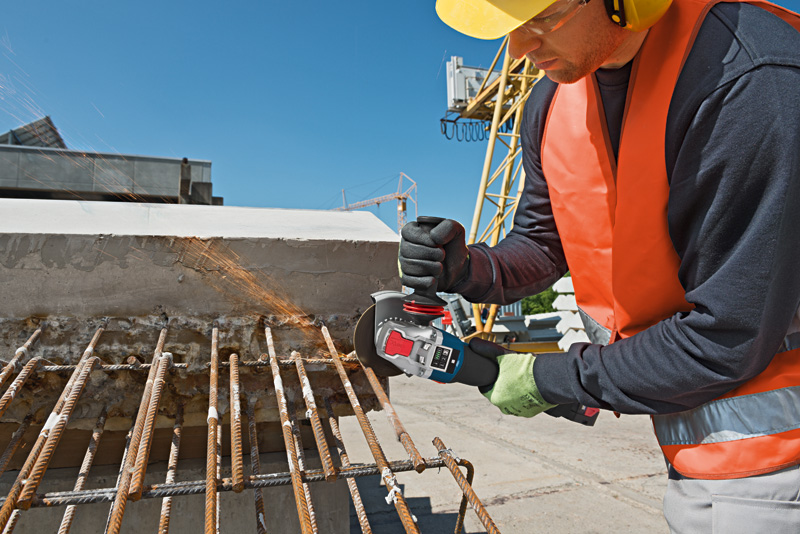 Eine Person mit Sicherheitsausrüstung verwendet einen Winkelschleifer, um auf einer Baustelle verrostete Bewehrungsstäbe zu schneiden. Anwendungsbild