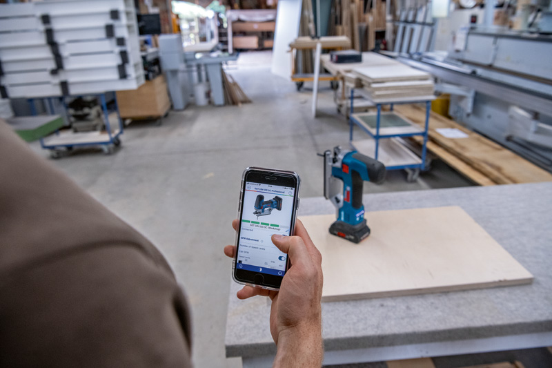 Eine Person verwendet ein Smartphone, um in einer Holzwerkstatt auf eine Akkubohrmaschine zu verweisen. Anwendungsbild
