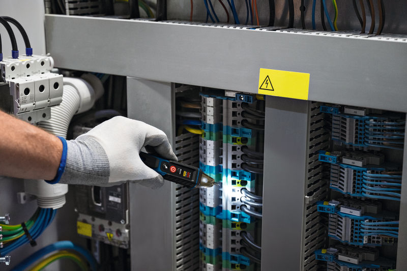 Eine Person mit Sicherheitsausrüstung überprüft mit einem Spannungsprüfer die elektrische Verkabelung in einem Bedienfeld. Anwendungsbild