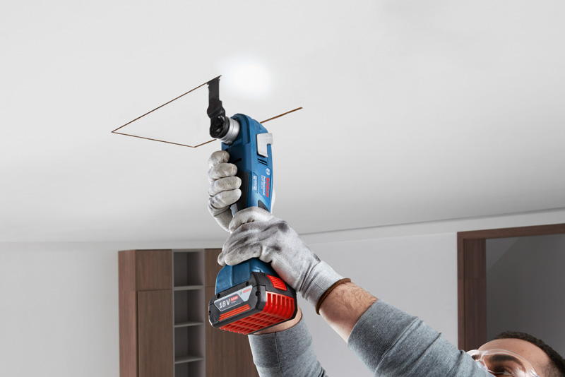 Eine Person mit Schutzausrüstung schneidet mit einem Akku-Elektrowerkzeug eine quadratische Öffnung in die Decke. Anwendungsbild Nahaufnahme