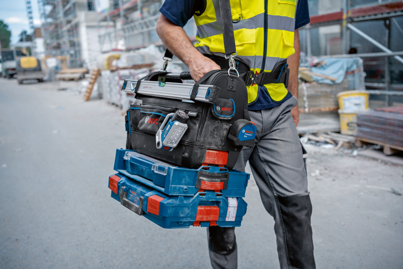 Eine Person mit Sicherheitsausrüstung trägt auf einer Baustelle gestapelte Werkzeugkisten und einen digitalen Winkelmesser. Anwendungsbild
