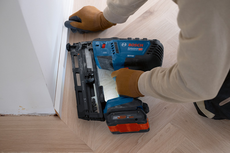 Eine Person mit Sicherheitsausrüstung befestigt mit einem Akku-Holznagler eine Zierleiste am Boden. Anwendungsbild