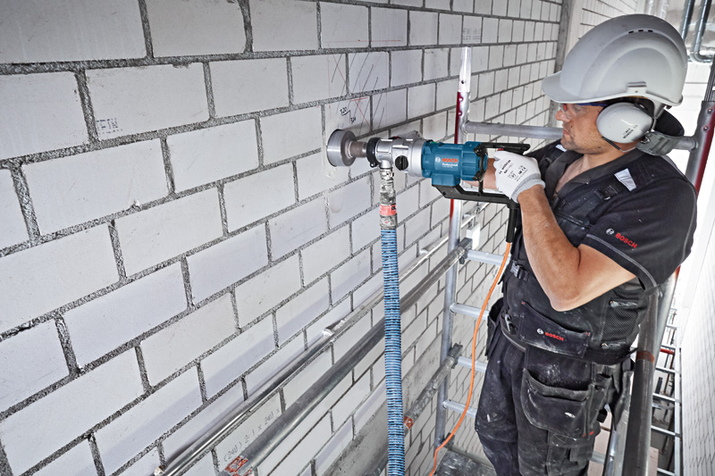 Eine Person mit Schutzausrüstung bohrt mit einem Diamantbohrer in eine Ziegelwand. Anwendungsbild
