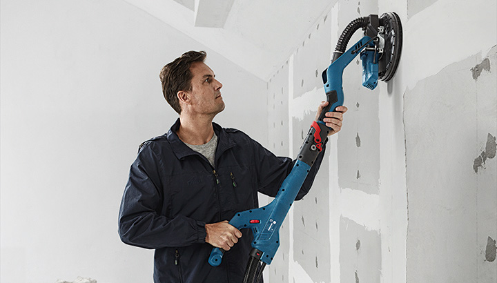 Eine Person schleift mit einem elektrischen Trockenbauschleifer Trockenbauwände an einer unfertigen Wand. Anwendungsbild