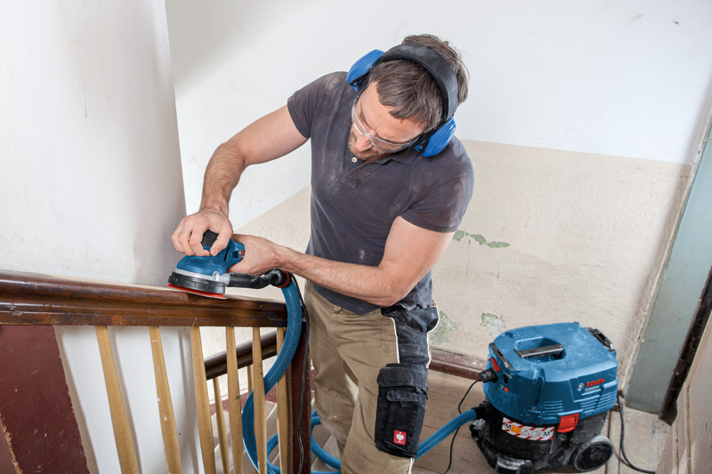 Eine Person mit Schutzausrüstung schleift einen Holzhandlauf mit einem Exzenterschleifer. Anwendungsbild