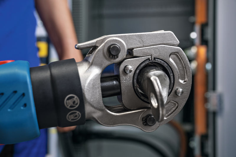 Eine Person mit Sicherheitsausrüstung bedient ein kabelloses hydraulisches Crimpwerkzeug vor einer Platte. Anwendungsbild