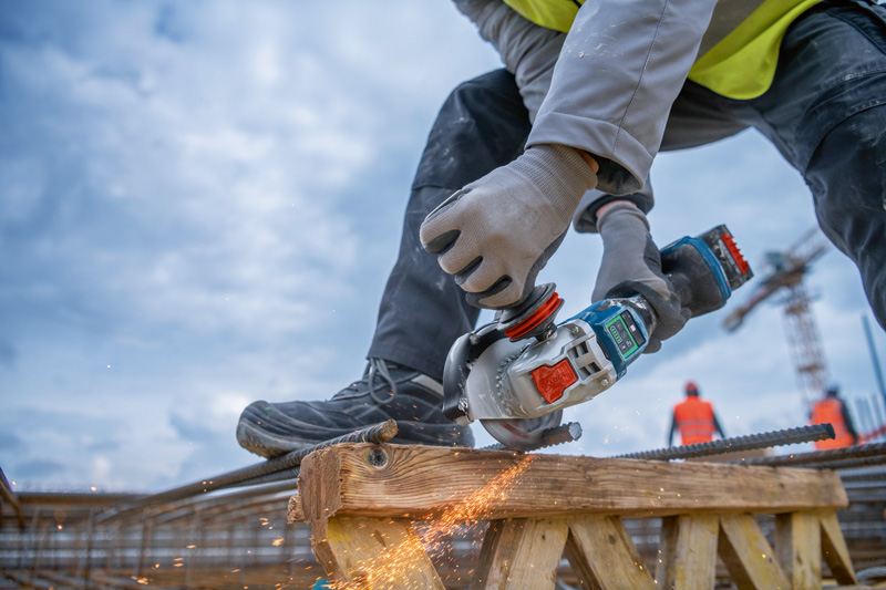 Eine Person mit Sicherheitsausrüstung schleift auf einer Baustelle Bewehrungsstahl mit einem Akku-Winkelschleifer. Anwendungsbild
