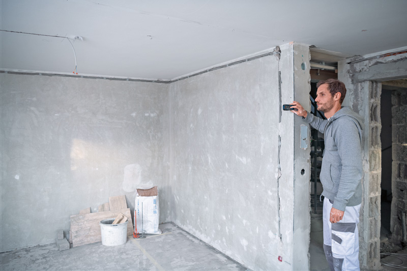 Eine Person misst mit einem Lasermessgerät eine Wand in einem leeren Raum. Anwendungsbild
