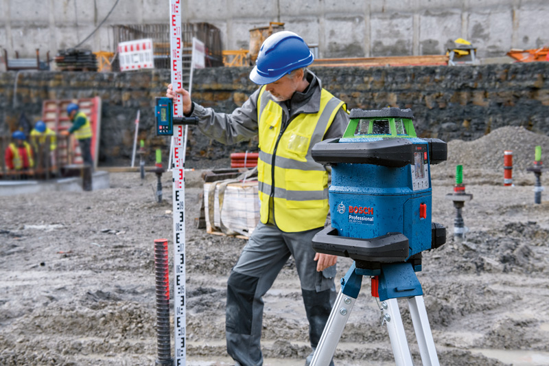 Eine Person mit Sicherheitsausrüstung verwendet auf einer schlammigen Baustelle ein Lasernivelliergerät und einen Empfänger. Anwendungsbild