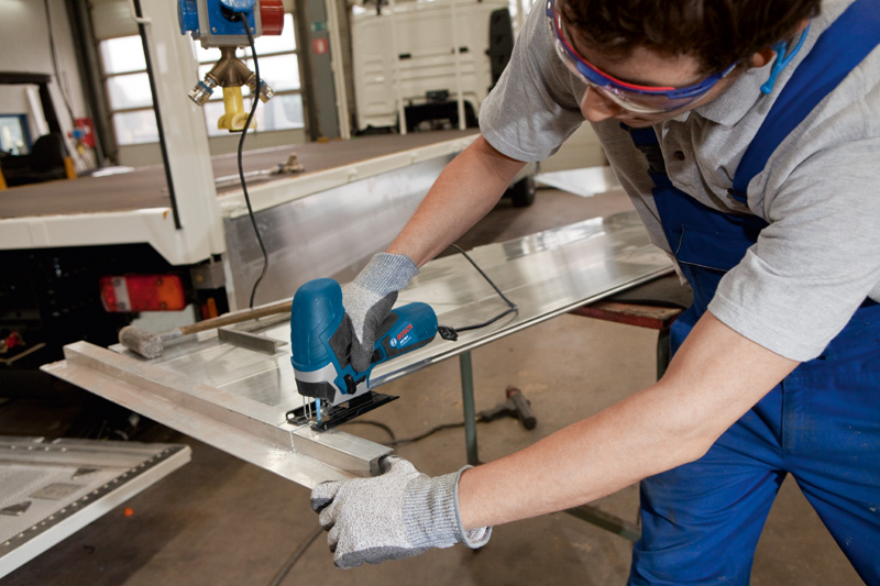 Eine Person mit Schutzausrüstung schneidet mit einer Stichsäge ein Metallblech. Anwendungsbild